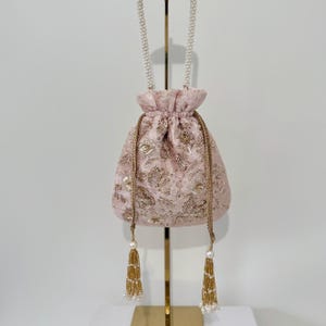 Puede incluir: Un bolso rosa con cordón y bordado floral dorado y una correa de cuentas de perlas. El bolso tiene borlas doradas con detalles de perlas. El bolso se exhibe en un soporte dorado.