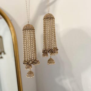 Könnte beinhalten: Ein Paar goldene und weiße Perlen-Brautschmuckstücke namens Kalire, die an einer weißen Wand hängen. Die Kalire bestehen aus goldenen Ketten mit kleinen weißen Perlen und haben unten eine glockenförmige Form.