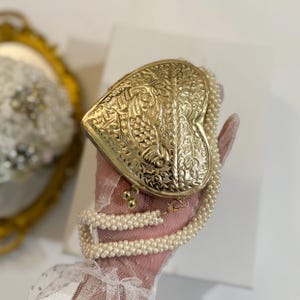 May include: A heart-shaped, gold-colored clutch with intricate floral and leaf patterns. The clutch has a pearl beaded strap. The clasp is gold-colored. The background is white.