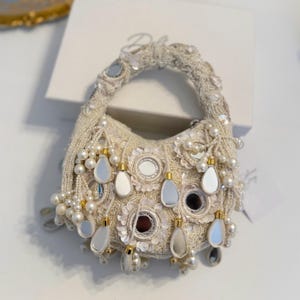 Può includere: Una borsa a mano bianca con perline, con un manico rotondo e una frangia di perle e dettagli a specchio.