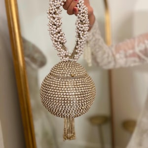 Op de afbeelding: Een goudkleurige, met kralen bezette handtas met een ronde body en een met kralen bezette handgreep. De tas is bedekt met kleine, glinsterende kralen en heeft een kwast aan de onderkant. De handgreep is gemaakt van witte parels.