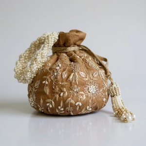 May include: A tan drawstring pouch bag featuring intricate white and gold floral embroidery. The bag includes a braided handle made of small white beads and a matching decorative tassel with white and gold beads hanging from the side drawstring.