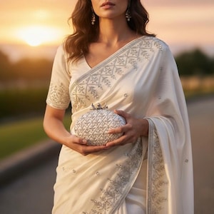 May include: Ivory sari with detailed silver embroidery and a matching blouse. A silver, patterned clutch is held in front. The background shows a sunset. The sari is draped.