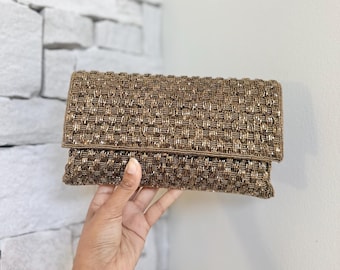 May include: A rectangular, beaded clutch in shades of brown and gold. The clutch has a woven pattern and a fold-over flap. The bag is held by a hand, showcasing the texture and color of the beads. The background is a white brick wall.
