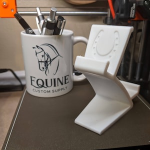 May include: White ceramic mug with a black horse design and the text "EQUINE CUSTOM SUPPLY". A white 3D printed phone stand with a horseshoe design is next to the mug.
