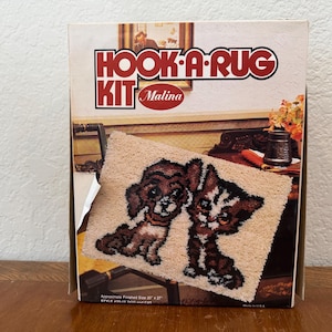May include: A "Hook-A-Rug Kit" box featuring a finished rug with two cartoon kittens. The rug is beige with brown and black details. The box has the text "HOOK-A-RUG KIT Malina" in red and white.