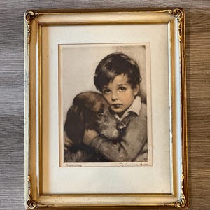 May include: Framed monochrome print of a child embracing a dog. The print is in a gold-coloured ornate frame. The child has dark hair and wears a sweater and collared shirt. The dog has long ears and is brown and black.