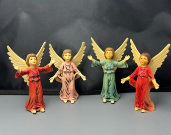 May include: Four small angel figurines with outstretched arms and cream-colored wings. The angels wear robes in red, pink, and green. The figurines are likely made of ceramic or a similar material, and are suitable for holiday or religious decor.