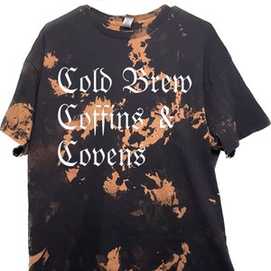 Pode incluir: Camiseta preta tie-dye com texto branco que diz "Cold Brew Coffins & Covens".