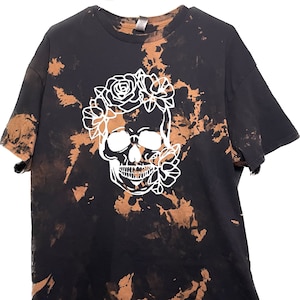 Puede incluir: Camiseta negra con un diseño blanqueado en blanco de una calavera con flores. La camiseta tiene un patrón tie-dye en tonos marrones y negros.