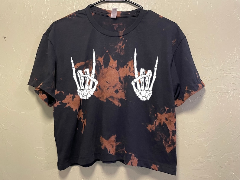 Puede incluir: Camiseta negra recortada con un patr&oacute;n de tie-dye y manos de esqueleto blancas haciendo el gesto de rock and roll.