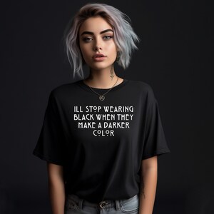 May include: A woman with short, silver hair is wearing a black t-shirt with the text "I'll stop wearing black when they make a darker color" printed in white.