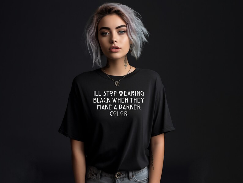 May include: A woman with short, silver hair is wearing a black t-shirt with the text "I'll stop wearing black when they make a darker color" printed in white.