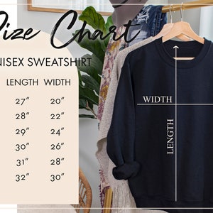 May include: Size chart for a unisex sweatshirt. The chart shows the length and width in inches for sizes S, M, L, XL, 2X, and 3X. A black sweatshirt is hanging on a wooden hanger.