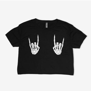 Puede incluir: Camiseta negra recortada con una mano de esqueleto blanca haciendo el signo de rock and roll a cada lado de la camiseta.
