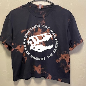 May include: Black t-shirt with a white dinosaur skull graphic inside a circle with the text "DINOSAURS EAT MAN WOMAN INHERITS THE EARTH". The shirt has a bleached, distressed design.