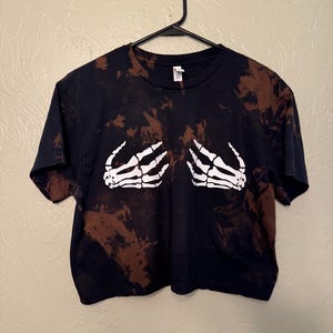 Może przedstawiać: Kr&oacute;tki t-shirt w kolorze czarnym i brązowym tie-dye z białym nadrukiem szkieletu dłoni.