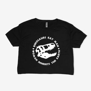 May include: Black crop top with a white dinosaur skull graphic inside a circular text design. The text reads "DINOSAURS EAT MAN WOMAN INHERITS THE EARTH".