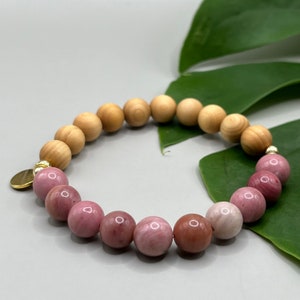 Op de afbeelding: Een roze en houten kralen armband met een gouden bedel. De armband is gemaakt van natuurlijke stenen en houten kralen.