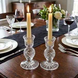 May include: Two clear glass candle holders with a faceted, cut-glass pattern and a wide, circular base. Each holder contains a single, lit, cream-colored taper candle. The holders are positioned on a dark wooden table with a black runner.