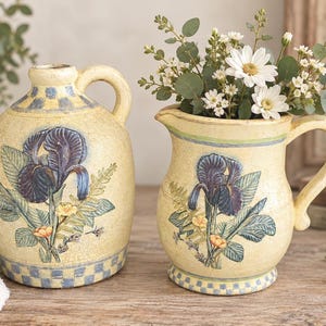 May include: A cream-colored ceramic jug and pitcher set featuring a hand-painted dark purple iris flower design with green leaves and yellow accents. Both pieces have a blue and cream checkered pattern at the base and a textured, crackled finish.