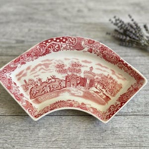 May include: A fan-shaped ceramic dish featuring a red and white transferware pattern. The design depicts a landscape with a stone structure, trees, and figures, surrounded by a decorative floral border along the curved edges of the dish.