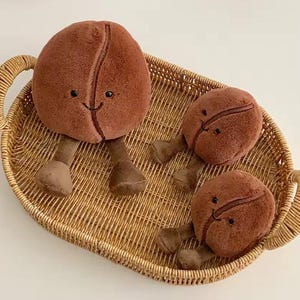 Puede incluir: Tres juguetes de peluche con forma de grano de caf&eacute; con caras sonrientes y patas peque&ntilde;as est&aacute;n colocados en una bandeja de mimbre tejida. Los granos de caf&eacute; son de color marr&oacute;n c&aacute;lido y la bandeja tiene un tono marr&oacute;n claro natural.