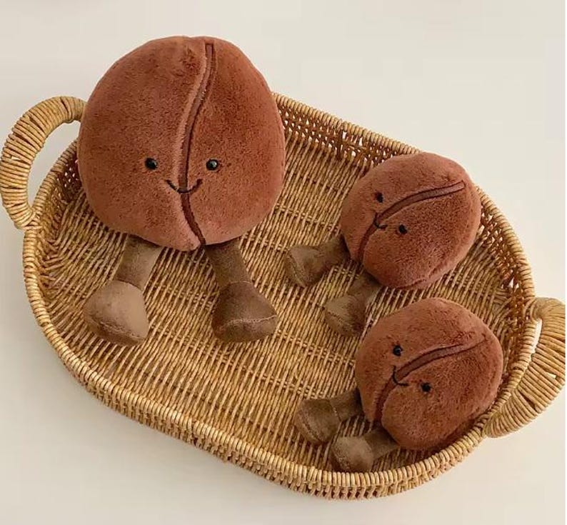 Puede incluir: Tres juguetes de peluche con forma de grano de caf&eacute; con caras sonrientes y patas peque&ntilde;as est&aacute;n colocados en una bandeja de mimbre tejida. Los granos de caf&eacute; son de color marr&oacute;n c&aacute;lido y la bandeja tiene un tono marr&oacute;n claro natural.