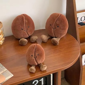 Puede incluir: Tres juguetes de peluche con forma de grano de caf&eacute; de diferentes tama&ntilde;os est&aacute;n sobre una superficie de madera. Cada grano es de color marr&oacute;n, con una sonrisa cosida y peque&ntilde;as piernas. Los juguetes est&aacute;n colocados sobre una mesa redonda de madera, con un calendario y un tarro de aperitivos en el fondo.