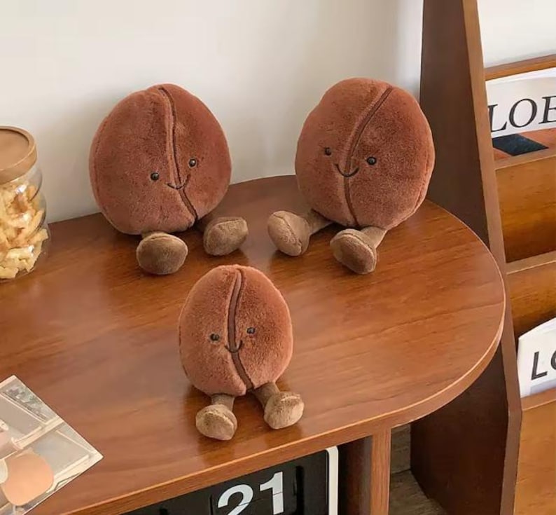 Puede incluir: Tres juguetes de peluche con forma de grano de caf&eacute; de diferentes tama&ntilde;os est&aacute;n sobre una superficie de madera. Cada grano es de color marr&oacute;n, con una sonrisa cosida y peque&ntilde;as piernas. Los juguetes est&aacute;n colocados sobre una mesa redonda de madera, con un calendario y un tarro de aperitivos en el fondo.
