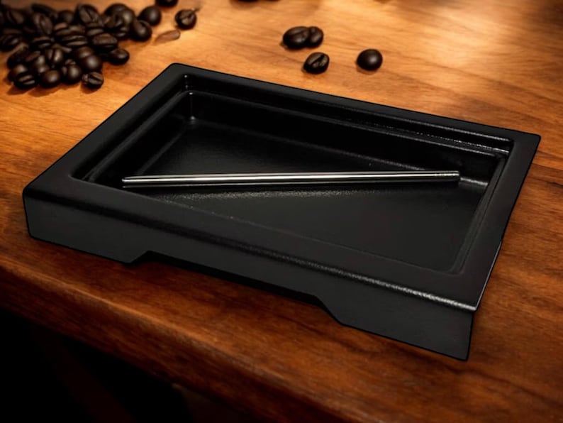 May include: A black rectangular tray with a raised edge and a small metal rod inside. The tray is sitting on a wooden surface.