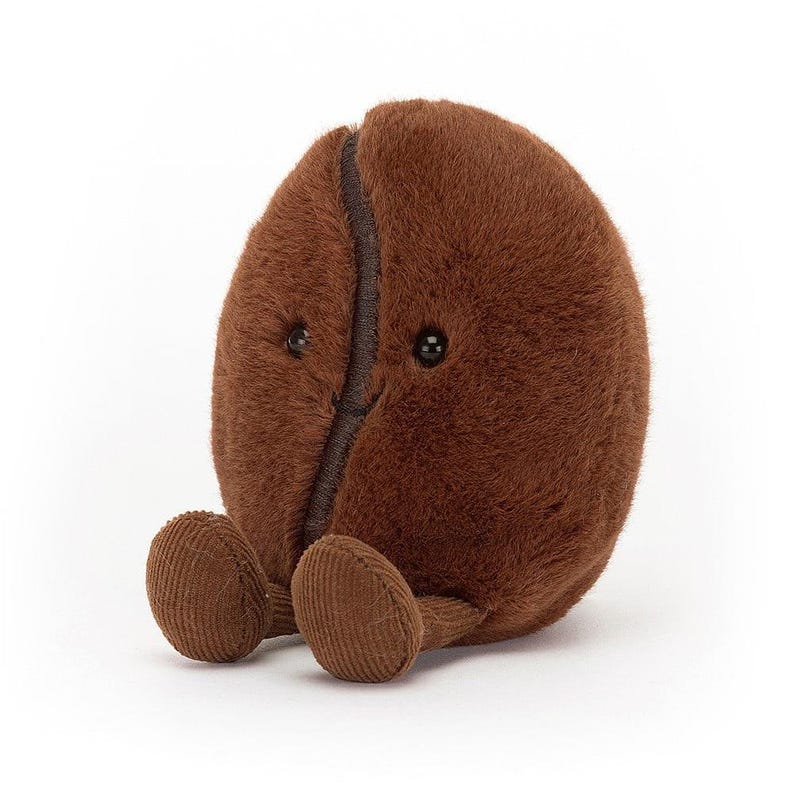 Puede incluir: Un juguete de peluche con forma de grano de caf&eacute; marr&oacute;n con una superficie suave y texturizada. El grano tiene peque&ntilde;os ojos negros de bot&oacute;n y pies de pana. El juguete est&aacute; dise&ntilde;ado para sentarse erguido y tiene una costura marr&oacute;n oscuro en el centro.