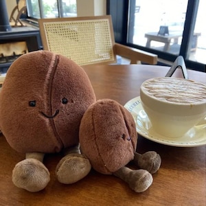 Puede incluir: Dos juguetes de peluche con forma de grano de caf&eacute; marr&oacute;n con caras sonrientes y patas peque&ntilde;as est&aacute;n sobre una mesa de madera. Una taza de caf&eacute; con arte latte est&aacute; en el fondo. La taza de caf&eacute; es blanca.
