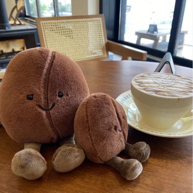 Puede incluir: Dos juguetes de peluche con forma de grano de caf&eacute; marr&oacute;n con caras sonrientes y patas peque&ntilde;as est&aacute;n sobre una mesa de madera. Una taza de caf&eacute; con arte latte est&aacute; en el fondo. La taza de caf&eacute; es blanca.