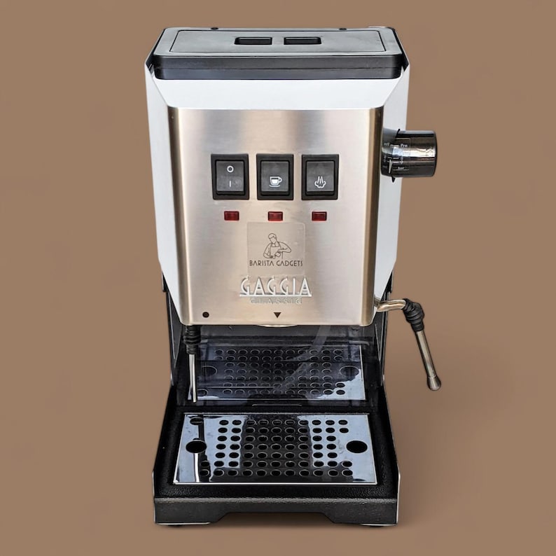 May include: A white and stainless steel espresso machine with a black drip tray. The machine has three buttons with icons for single, double, and hot water. The machine has the text "Barista Gadgets" and "Gaggia" on the front.