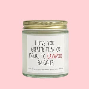 Puede incluir: Una vela de vidrio en un frasco con tapa dorada y una etiqueta blanca que dice "I love you greater than or equal to cavapoo snuggles". La etiqueta también incluye el texto "KWB | Fragrant and Long Lasting Apricot Coconut Wax".