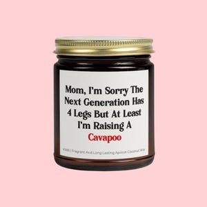 Puede incluir: Una vela de vidrio marrón en un frasco con tapa dorada. La etiqueta dice "Mamá, lo siento, la próxima generación tiene 4 patas, pero al menos estoy criando un Cavapoo". La parte inferior de la etiqueta dice "KWB | Cera de coco y albaricoque fragante y de larga duración".