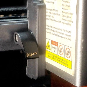 May include: Close-up of a black laser engraver with a silver label that reads "LASER APERTURE" and includes safety instructions. The label also includes the company name, address, and contact information. The laser engraver has a black switch with the word "Viper" printed on it.