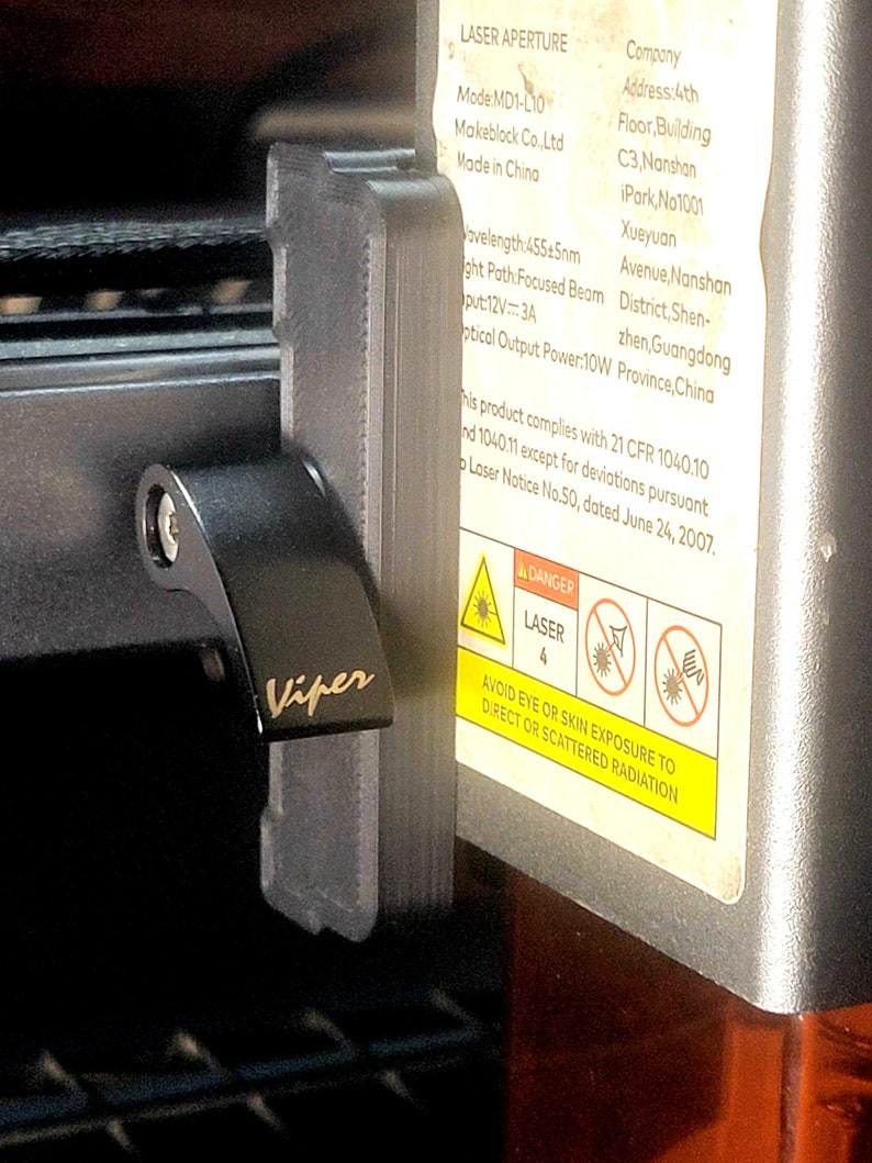 May include: Close-up of a black laser engraver with a silver label that reads "LASER APERTURE" and includes safety instructions. The label also includes the company name, address, and contact information. The laser engraver has a black switch with the word "Viper" printed on it.