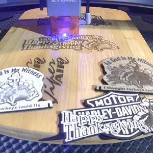 May include: A wooden cutting board with laser-etched designs, including Harley-Davidson logos and Thanksgiving-themed graphics. The board also features the text "As God Is My Witness" and "I thought turkeys could fly..."