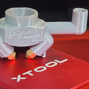 May include: A light gray 3D-printed part with the text "Viper Laser" on the front. The part has a cylindrical top, a rectangular body, and two orange and gold-colored nozzles. It is placed on a red surface with the text "XTOOL".