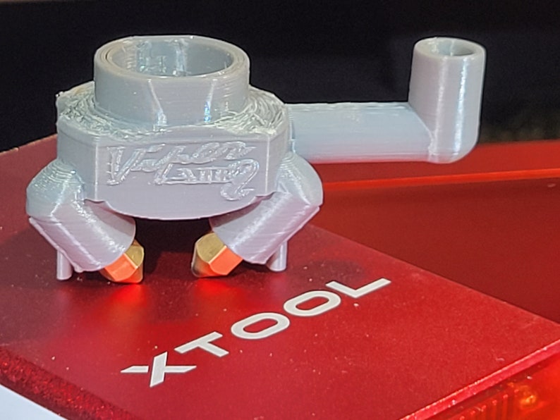 May include: A light gray 3D-printed part with the text "Viper Laser" on the front. The part has a cylindrical top, a rectangular body, and two orange and gold-colored nozzles. It is placed on a red surface with the text "XTOOL".