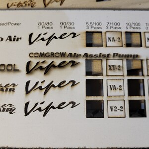 May include: A light-colored wooden panel with laser-etched text and squares. The text includes "Speed/Power", "No Air", "COMGROW Air Assist Pump", "XTOOL", and "Viper". The squares contain etched patterns and labels like "NA-2" and "XT-2".