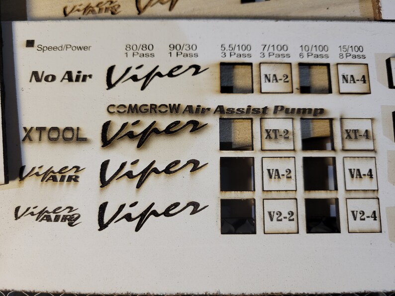 May include: A light-colored wooden panel with laser-etched text and squares. The text includes "Speed/Power", "No Air", "COMGROW Air Assist Pump", "XTOOL", and "Viper". The squares contain etched patterns and labels like "NA-2" and "XT-2".