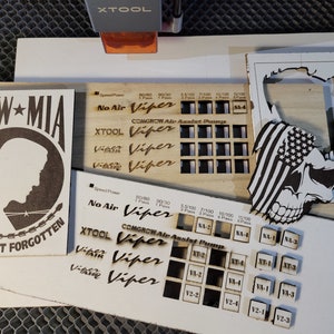 May include: Laser-cut wooden designs, including a POW-MIA tribute with the text "YOU ARE NOT FORGOTTEN", a skull with an American flag, and various test patterns with the "XTOOL Viper" logo. The designs are on light-colored wood.