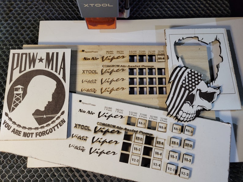 May include: Laser-cut wooden designs, including a POW-MIA tribute with the text "YOU ARE NOT FORGOTTEN", a skull with an American flag, and various test patterns with the "XTOOL Viper" logo. The designs are on light-colored wood.