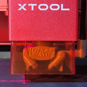 May include: Close-up of a red XTOOL laser engraving machine. The machine's housing is red with the word "XTOOL" in white. The laser is focused on a small, tan-colored object with the text "Viper Air?" engraved on it. A pink laser beam is visible.