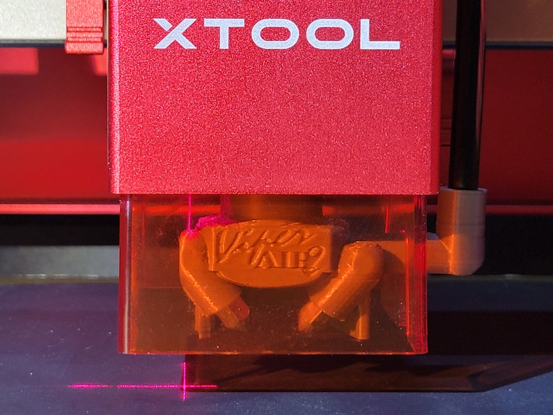 May include: Close-up of a red XTOOL laser engraving machine. The machine's housing is red with the word "XTOOL" in white. The laser is focused on a small, tan-colored object with the text "Viper Air?" engraved on it. A pink laser beam is visible.
