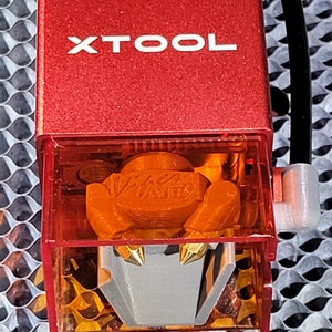 May include: Close-up of a red XTOOL laser engraver head. The clear plastic housing reveals an orange interior with a silver and gold nozzle. A black cable and white connector are attached to the side.