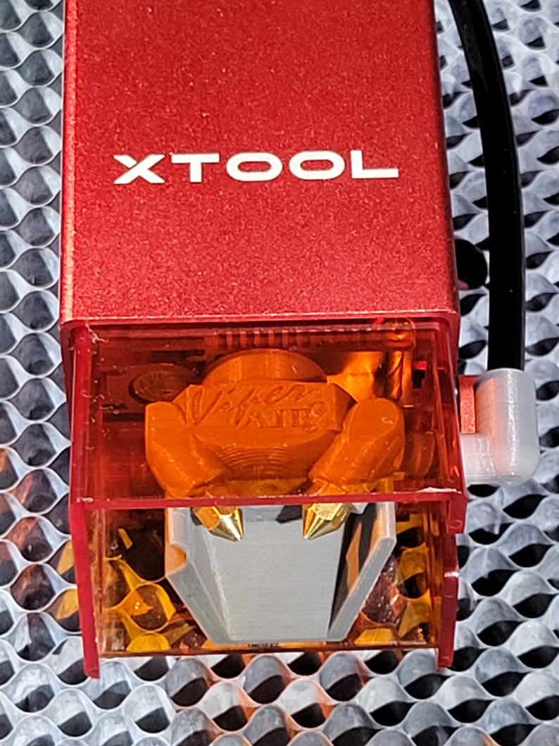 May include: Close-up of a red XTOOL laser engraver head. The clear plastic housing reveals an orange interior with a silver and gold nozzle. A black cable and white connector are attached to the side.